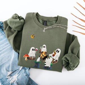 May include: Olive green sweatshirt with a graphic of cartoon ghosts around a campfire, drinking wine and eating popcorn. A silver necklace is draped on the sweatshirt. A pair of blue jeans is in the background.