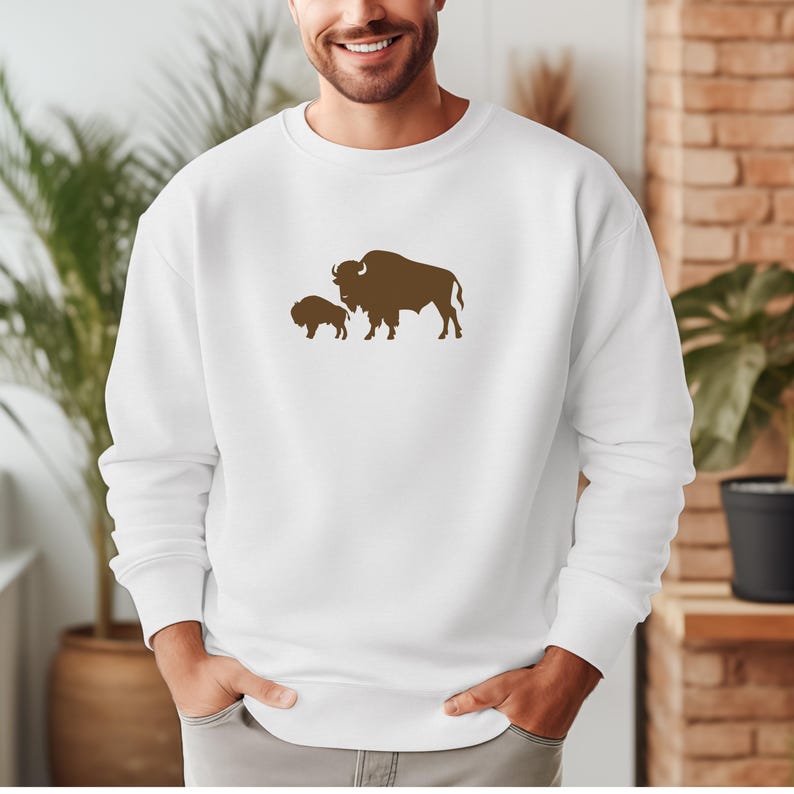 May include: A white long-sleeve crewneck sweatshirt featuring a brown graphic print of a large adult bison standing next to a smaller bison calf centred on the chest. The garment has a ribbed neckline, cuffs, and hem, providing a clean look.