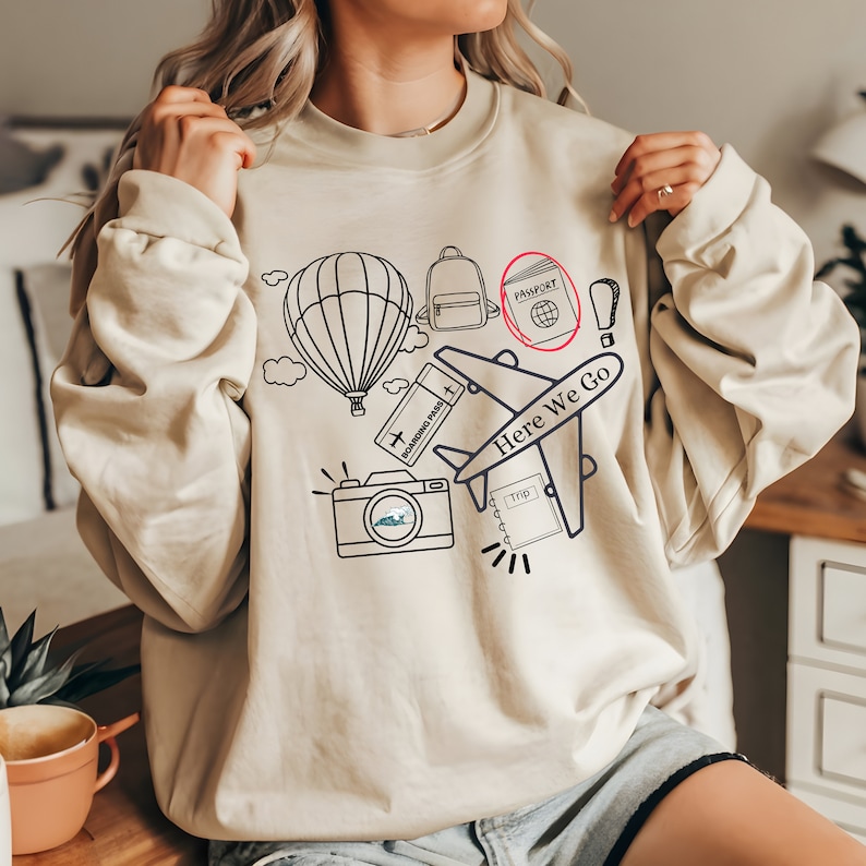 May include: A beige sweatshirt with a graphic design of travel icons, including a hot air balloon, a backpack, a passport, a plane, a camera, and a boarding pass. The text "Here We Go" is printed on the sweatshirt.