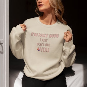 May include: A beige sweatshirt with the text 'I'M NOT SHY i JUST DON'T LIKE YOU' in pink lettering. The text is stylized with a pink outline. A pink middle finger emoji is next to the word 'YOU'.