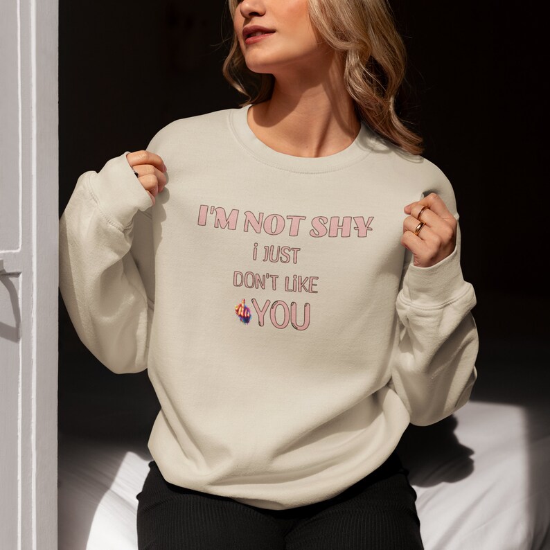 May include: A beige sweatshirt with the text 'I'M NOT SHY i JUST DON'T LIKE YOU' in pink lettering. The text is stylized with a pink outline. A pink middle finger emoji is next to the word 'YOU'.