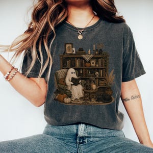 Könnte beinhalten: Ein anthrazitfarbenes T-Shirt mit einem Grafikdruck eines weißen Geistes, der in einem Sessel sitzt und ein Buch liest. Die Szene zeigt ein hölzernes Bücherregal mit Büchern, Kürbissen, Kerzen, einem Totenkopf und einer schwarzen Katze auf einem Teppich.