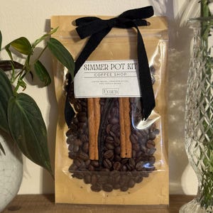 May include: A cellophane bag containing coffee beans, cinnamon sticks, and vanilla beans, tied with a black ribbon. The label reads "Simmer Pot Kit Coffee Shop". The bag is presented on a wooden surface, suggesting a rustic aesthetic.
