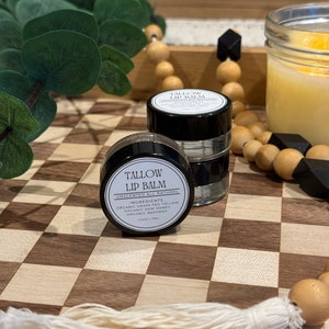 May include: A small black jar of unscented tallow lip balm with the label "Tallow Lip Balm" and the ingredients listed: "Organic Grass Fed Tallow, Organic Raw Honey, Organic Beeswax". The jar is sitting on a wooden surface with a checkered pattern.