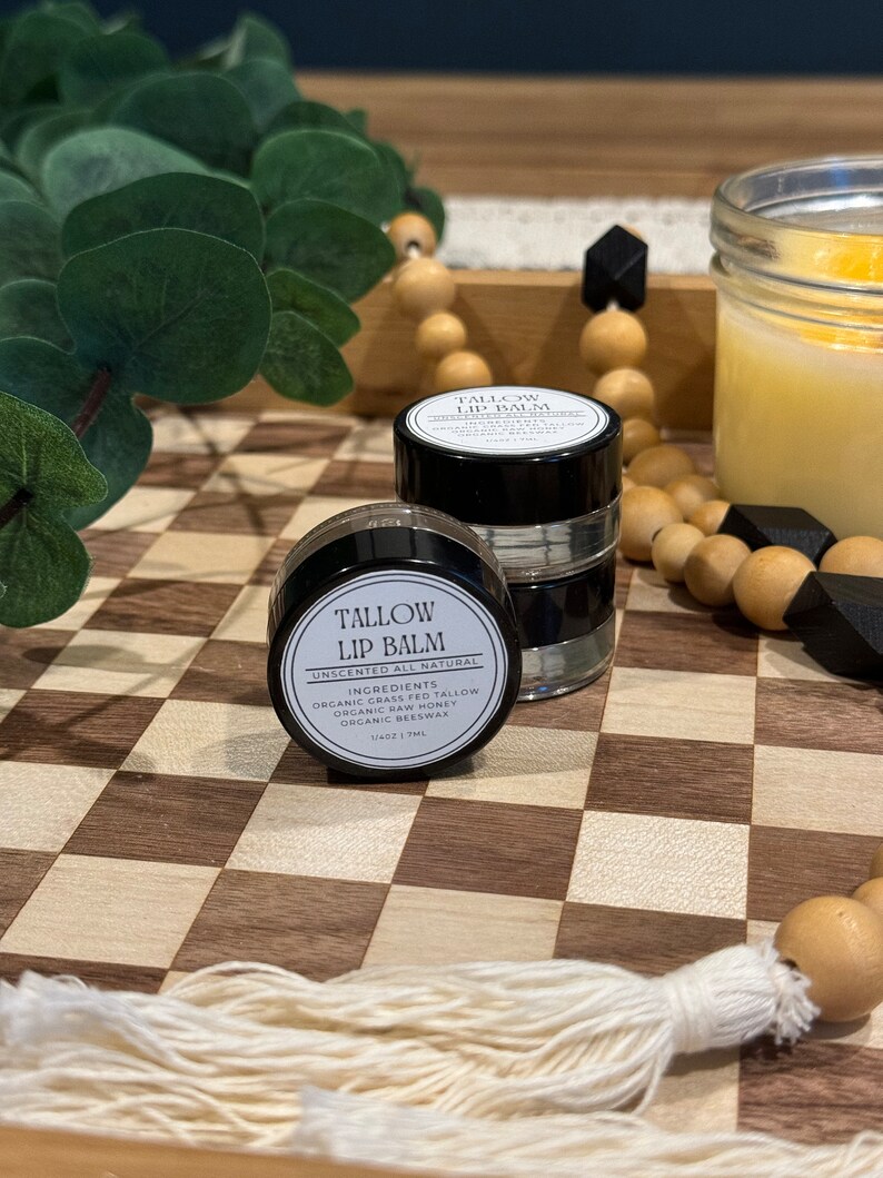 May include: A small black jar of unscented tallow lip balm with the label "Tallow Lip Balm" and the ingredients listed: "Organic Grass Fed Tallow, Organic Raw Honey, Organic Beeswax". The jar is sitting on a wooden surface with a checkered pattern.