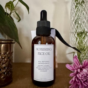 May include: A brown glass bottle of Nourishing Face Oil with a black dropper and label. The label lists ingredients like organic jojoba, rose, and lavender. The bottle is 2 oz / 59ml. A purple flower and greenery are in the background.