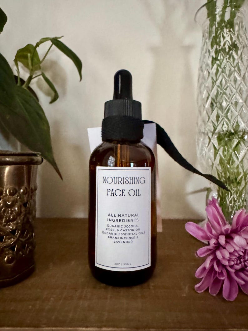 May include: A brown glass bottle of Nourishing Face Oil with a black dropper and label. The label lists ingredients like organic jojoba, rose, and lavender. The bottle is 2 oz / 59ml. A purple flower and greenery are in the background.
