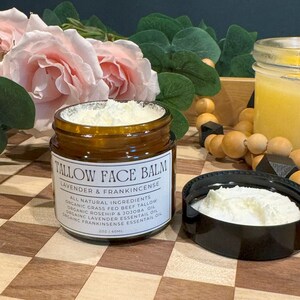 May include: A brown glass jar with a white lid filled with a white cream. The jar is labeled "Tallow Face Balm Lavender & Frankincense" and lists all natural ingredients. The jar is sitting on a wooden surface with a checkered pattern.