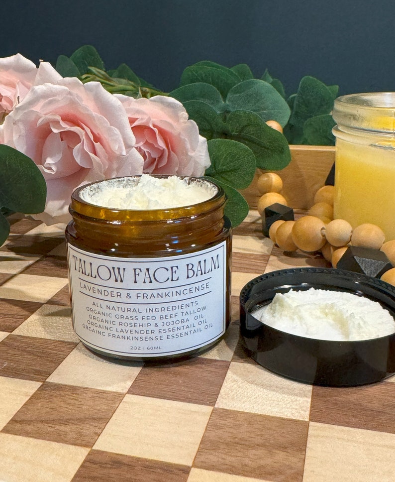 May include: A brown glass jar with a white lid filled with a white cream. The jar is labeled "Tallow Face Balm Lavender & Frankincense" and lists all natural ingredients. The jar is sitting on a wooden surface with a checkered pattern.