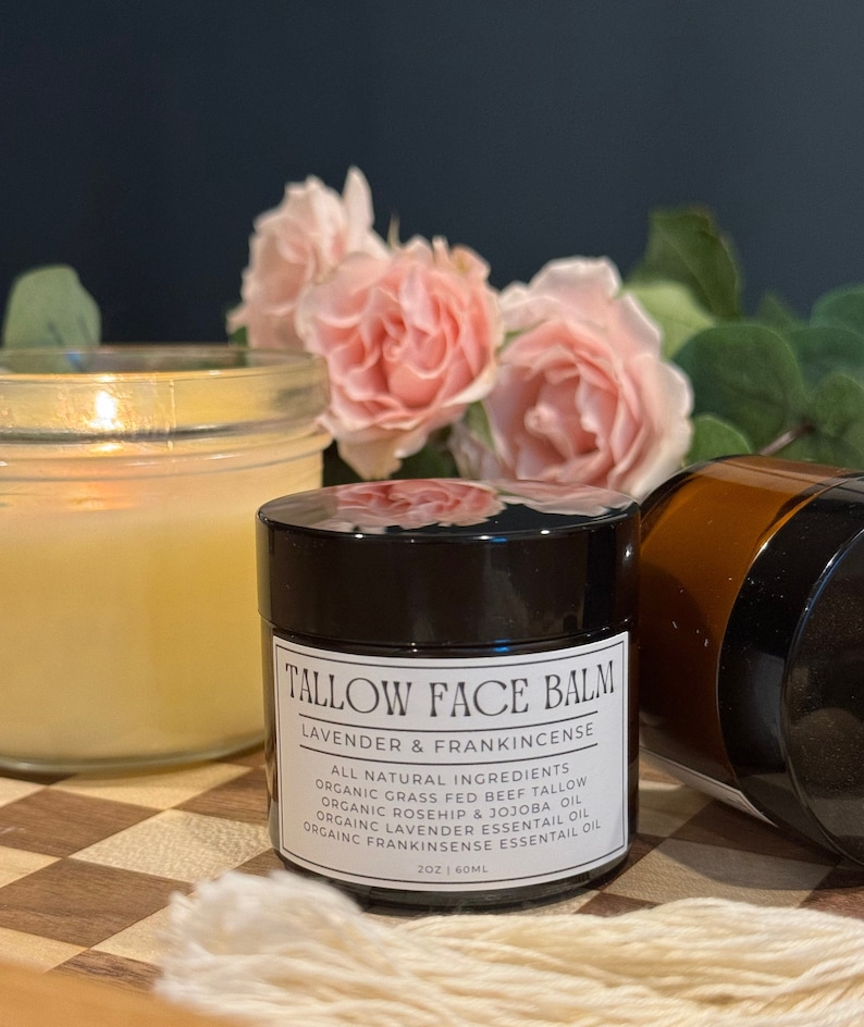 May include: A black jar of Tallow Face Balm with a white label that reads "Tallow Face Balm Lavender & Frankincense" and lists all natural ingredients. The jar is sitting on a checkered surface with a candle and a brown jar in the background.