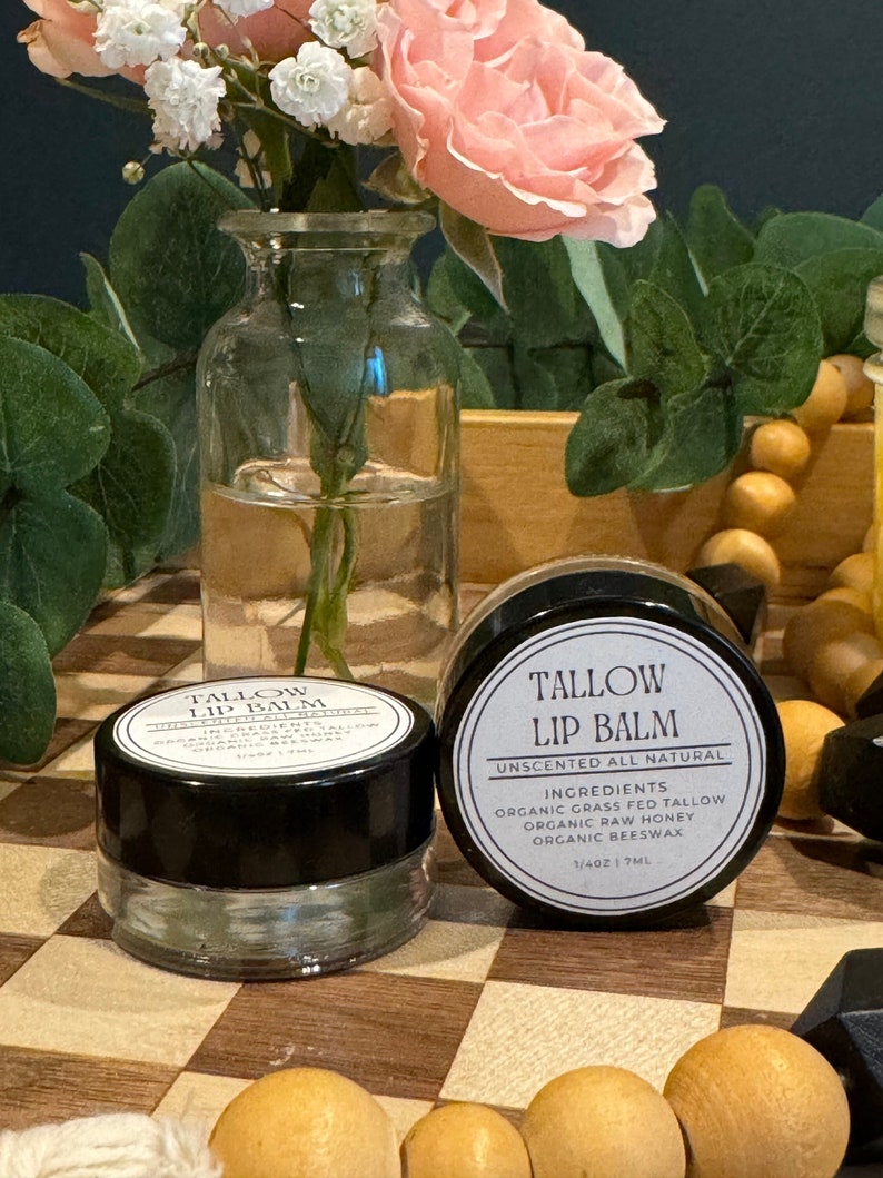 May include: Two small glass jars of lip balm with black lids. The jar on the right has a white label that reads "Tallow Lip Balm Unscented All Natural Ingredients Organic Grass Fed Tallow Organic Raw Honey Organic Beeswax 1/4oz 17ML".
