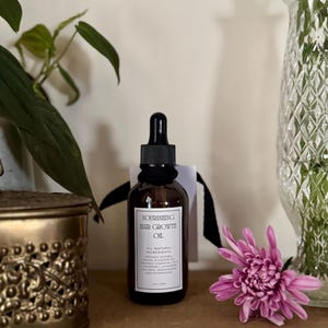 May include: A brown glass bottle of "Nourishing Hair Growth Oil" with a black dropper, a white label, and a black ribbon. The label lists all-natural ingredients. A pink flower and a decorative vase with flowers are in the background.