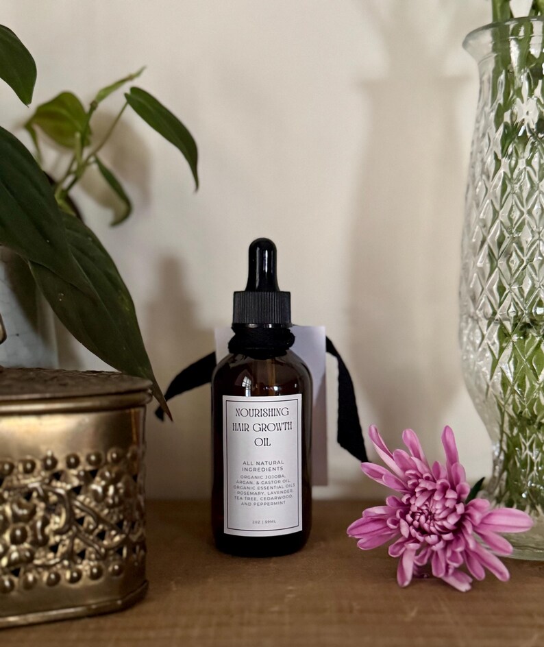 May include: A brown glass bottle of "Nourishing Hair Growth Oil" with a black dropper, a white label, and a black ribbon. The label lists all-natural ingredients. A pink flower and a decorative vase with flowers are in the background.
