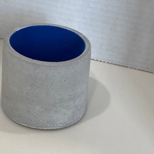 Puede incluir: Un pequeño macetero redondo de hormigón con un interior azul. El macetero está hecho de un material rugoso y texturizado y tiene un borde liso y redondeado.