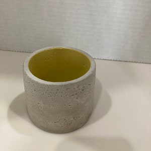 以下が含まれることがあります： 黄色い内側を持つ、小さく丸い白いコンクリート製のプランター。プランターはざらざらとした質感の表面をしています。