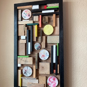 May include: A framed abstract art piece made of wood and painted circles. The piece is made up of various shapes and sizes of wood, some painted in different colors. The piece is framed in black.