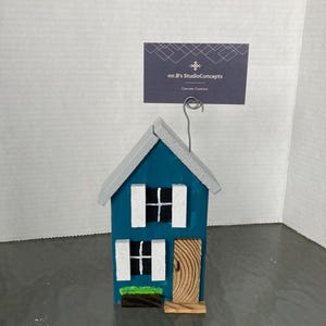May include: A teal blue wooden house with white shutters and a brown wooden door. The house has a green grass patch in front of it and a small wooden porch. The house is sitting on a white surface. A business card with the text "mr.B's StudioConcepts Concrete Creations" is attached to the top of the house.