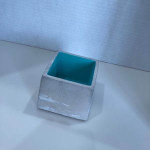 May include: A small, square concrete planter with a white, textured exterior and a bright turquoise interior.