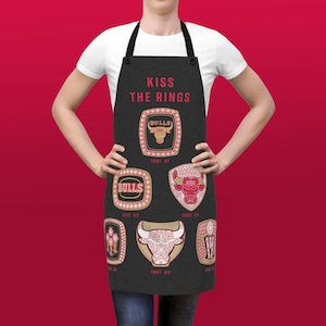 May include: Black apron featuring the text "Kiss the Rings" and images of six championship rings from the Chicago Bulls basketball team. Each ring displays the team logo and the year it was won.