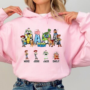 Może przedstawiać: R&oacute;żowa bluza z nadrukiem postaci z Toy Story, kt&oacute;re tworzą napis "MAMA" z nazwami "Mike, Jessi, Jack, Emily" pod spodem.