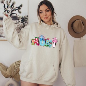 May include: A light beige hooded sweatshirt with a graphic print of the word "AUNT" with cartoon characters. The characters are Stitch, Lilo, and other Disney characters.