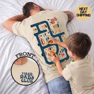 May include: Beige t-shirt with a blue road map design on the back, featuring colorful buildings and vehicles. The front of the shirt has a circular graphic with the text "TIRED DADS CLUB". A child plays with toy cars on the shirt.
