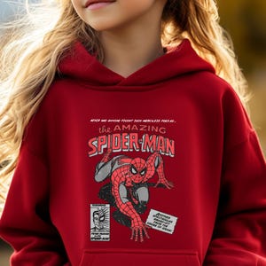May include: Red hooded sweatshirt featuring a vintage comic book style graphic of Spider-Man. The graphic reads "NEVER HAS ANYONE FOUGHT SUCH MERCILESS FOES AS... the AMAZING SPIDER-MAN".