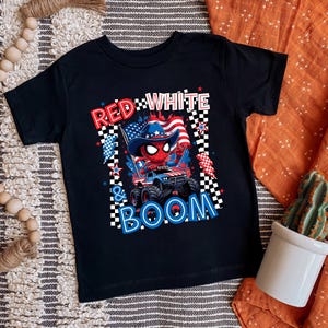 May include: Black t-shirt featuring a graphic of a Spider-Man-like character in a cowboy hat driving a monster truck with an American flag. The text reads "RED WHITE & BOOM" in red, white, and blue.