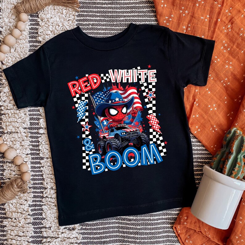 May include: Black t-shirt featuring a graphic of a Spider-Man-like character in a cowboy hat driving a monster truck with an American flag. The text reads "RED WHITE & BOOM" in red, white, and blue.