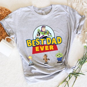 May include: A light gray t-shirt featuring the phrase "BEST DAD EVER" in yellow and blue block letters with a red banner. The shirt includes a cartoon image of Buzz Lightyear and other Toy Story characters: Bo Peep, Alex the horse, and Robert the alien.