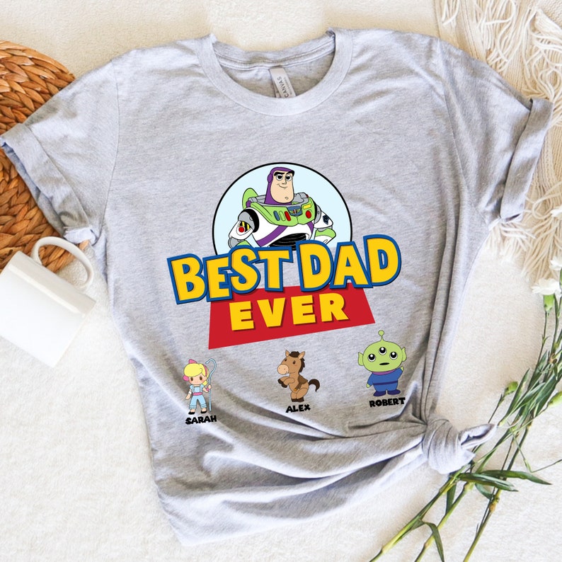 May include: A light gray t-shirt featuring the phrase "BEST DAD EVER" in yellow and blue block letters with a red banner. The shirt includes a cartoon image of Buzz Lightyear and other Toy Story characters: Bo Peep, Alex the horse, and Robert the alien.