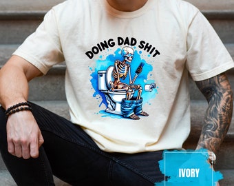 May include: A white t-shirt with a skeleton sitting on a toilet and the text "Doing Dad Shit" in blue and black lettering. The background is a blue watercolor design. The t-shirt is labeled "Ivory".