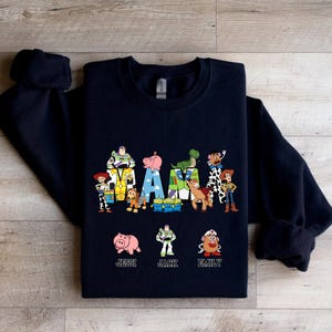 Może przedstawiać: Czarna bluza z motywem Toy Story. Słowo "MAMA" jest napisane postaciami z filmu. Pod słowem znajdują się imiona "Jessi", "Jack" i "Emily".