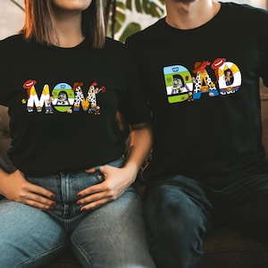 K&ouml;nnte beinhalten: Ein schwarzes T-Shirt mit dem Wort "MOM" in bunten Buchstaben, die Figuren aus dem Toy Story-Film-Franchise darstellen. Die Figuren sind Woody, Buzz Lightyear, Jessie und andere.