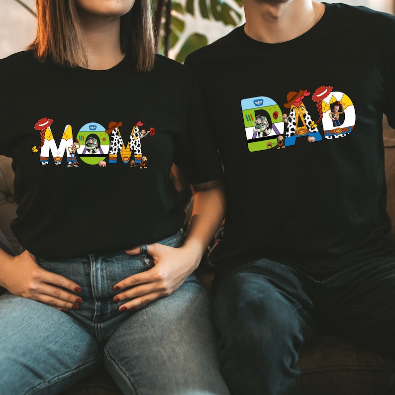 K&ouml;nnte beinhalten: Ein schwarzes T-Shirt mit dem Wort "MOM" in bunten Buchstaben, die Figuren aus dem Toy Story-Film-Franchise darstellen. Die Figuren sind Woody, Buzz Lightyear, Jessie und andere.