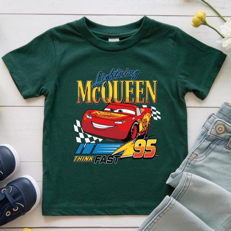 May include: A dark green t-shirt featuring a graphic of Lightning McQueen from the Cars movie. The design includes the text "Lightning McQueen", the number "95", and the phrase "Think Fast". The car is red with yellow accents.