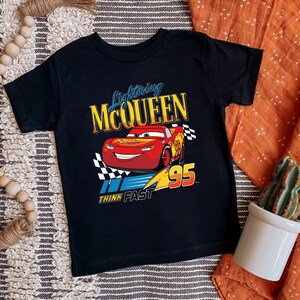 May include: Black t-shirt featuring Lightning McQueen from the Cars movie. The design includes a red race car with the number 95, a checkered flag, and the text "Lightning McQueen" and "Think Fast".