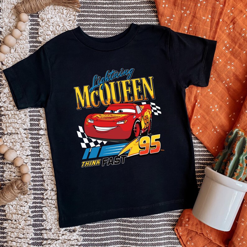 May include: Black t-shirt featuring Lightning McQueen from the Cars movie. The design includes a red race car with the number 95, a checkered flag, and the text "Lightning McQueen" and "Think Fast".