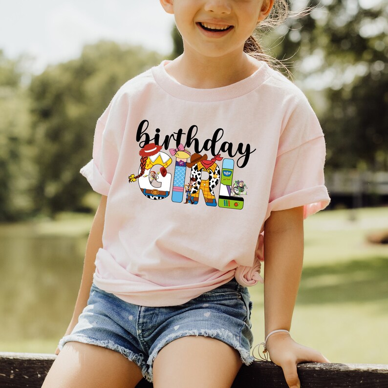 K&ouml;nnte beinhalten: Ein pinkfarbenes T-Shirt mit dem Schriftzug "birthday girl" in Schwarz. Die Buchstaben bestehen aus Figuren aus dem Toy Story-Film-Franchise.