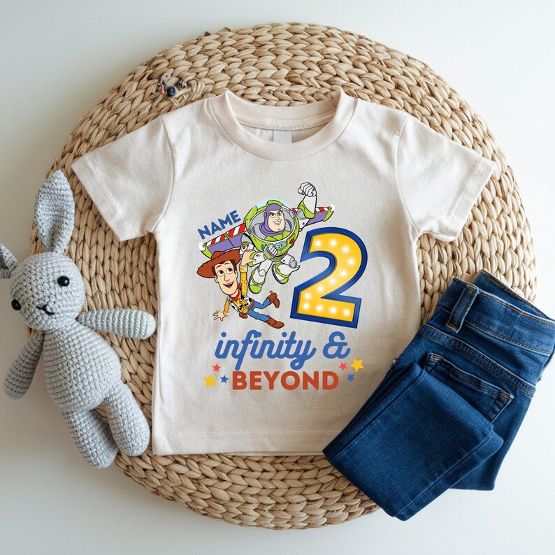K&ouml;nnte beinhalten: Ein cremefarbenes T-Shirt mit einer bunten Grafik von Buzz Lightyear und Woody aus Toy Story, mit der Zahl 2 und den Worten "infinity & beyond". Dazu eine blaue Jeans und ein graues Stoffkaninchen.