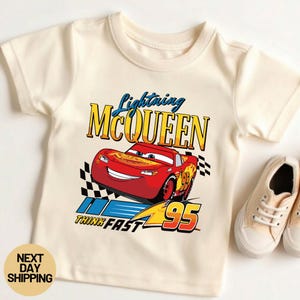 May include: Cream-colored t-shirt featuring a graphic of Lightning McQueen from the movie Cars. The design includes the text "Lightning McQueen", "Think Fast", and the number "95". A pair of beige sneakers are next to the shirt.