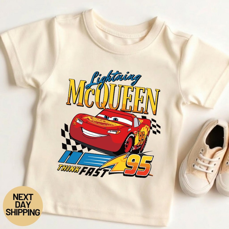 May include: Cream-colored t-shirt featuring a graphic of Lightning McQueen from the movie Cars. The design includes the text "Lightning McQueen", "Think Fast", and the number "95". A pair of beige sneakers are next to the shirt.