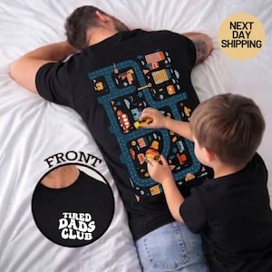May include: Black t-shirt with a road map design on the back, featuring buildings and vehicles. The front of the shirt has the text "TIRED DADS CLUB" in a circular design. A child is playing with toy cars on the shirt.