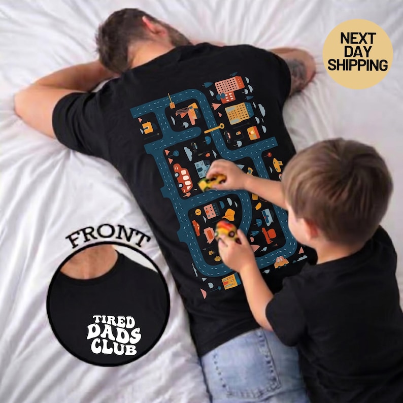 Pu&ograve; includere: T-shirt nero con un disegno di mappa stradale sul retro, con edifici e veicoli. La parte anteriore della maglietta presenta la scritta "TIRED DADS CLUB" in un design circolare. Un bambino gioca con macchinine sulla maglietta.