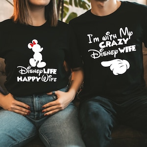 May include: Black t-shirts with white graphics. One shirt features a Mickey Mouse silhouette and the text "Disney Life Happy Wife." The other shirt says "I'm with My Crazy Disney Wife" with a pointing hand graphic. A fun couple's matching set.