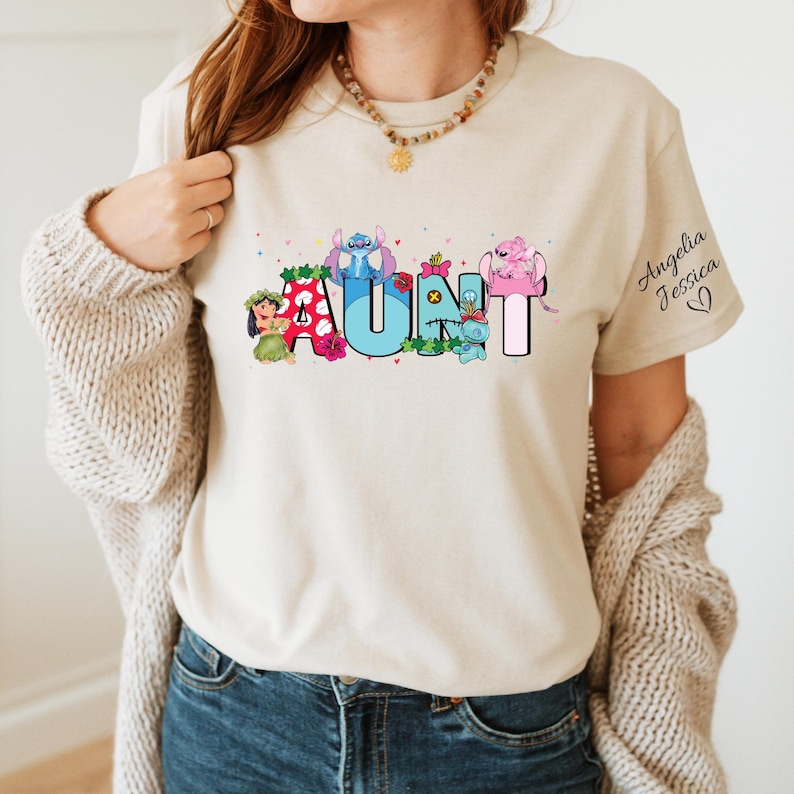 Puede incluir: Una camiseta beige con un gr&aacute;fico colorido de la palabra "AUNT" con personajes de dibujos animados de Lilo & Stitch y Moana de Disney. El texto "Angelia Jessica" est&aacute; escrito en cursiva en el lado derecho de la camiseta.