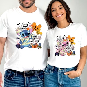 May include: White t-shirts featuring Halloween-themed cartoon characters. One shirt has a blue character wrapped in bandages, and the other has a pink character. Both shirts have spiderwebs, pumpkins, and ghosts.