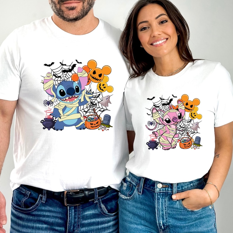 May include: White t-shirts featuring Halloween-themed cartoon characters. One shirt has a blue character wrapped in bandages, and the other has a pink character. Both shirts have spiderwebs, pumpkins, and ghosts.