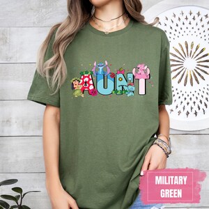 Puede incluir: Una camiseta verde con un gr&aacute;fico de personajes de Stitch que deletrean "AUNT" en letras de colores. La camiseta es de corte holgado y tiene cuello redondo.