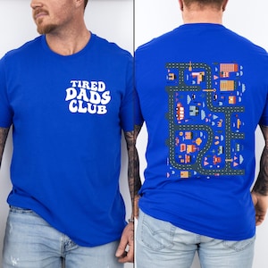 Puede incluir: Una camiseta azul real con el texto "TIRED DADS CLUB" en blanco en la parte delantera. La parte posterior de la camiseta presenta una ilustraci&oacute;n colorida de una ciudad con carreteras, edificios y coches.